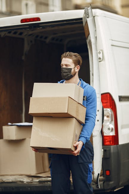 A man wearing a black face mask and a blue hoodie is standing outside a white moving van with its rear door open, preparing to load or unload cardboard boxes. He is holding three stacked boxes, includ