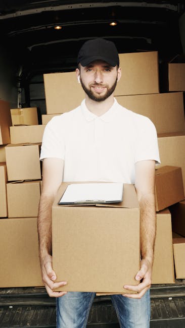 A man wearing a black face mask and a blue hoodie is standing outside a white moving van with its rear door open, preparing to load or unload cardboard boxes. He is holding three stacked boxes, includ