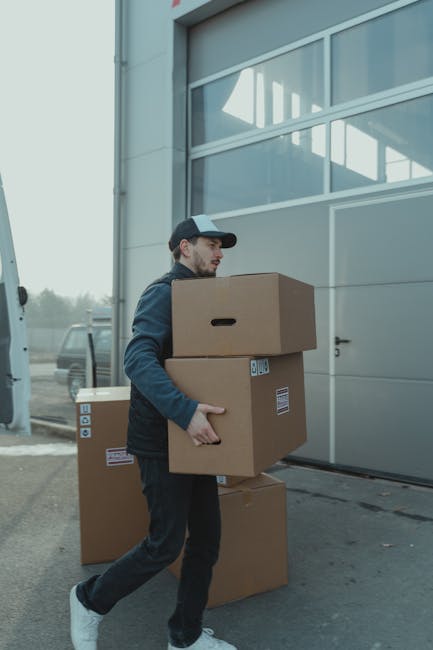 A man dressed in a blue hoodie and navy trousers, standing on a cobbled street near a white, large moving van with its rear doors open, revealing empty wooden-finished interior walls. He is holding an