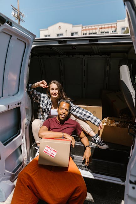 A professional removals worker with a friendly expression is standing outside a white moving van during a home relocation process. The worker, wearing a white polo shirt and a black cap, is carrying a