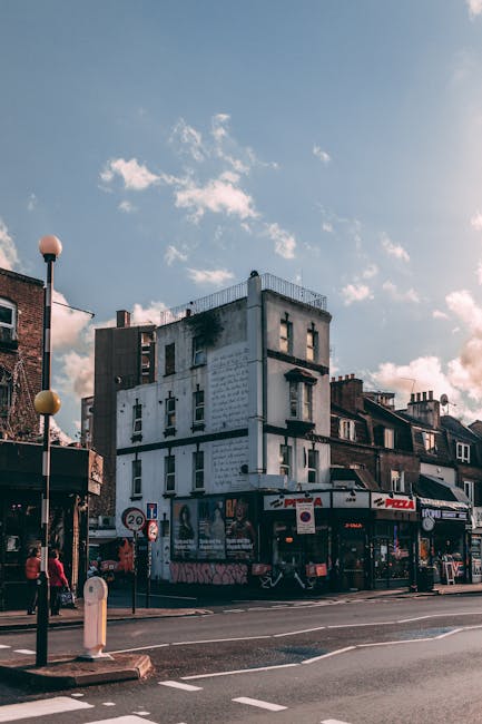 A multi-story building located on a busy urban street corner, featuring a white facade with black horizontal lines and several windows, some of which appear partially boarded up or covered. The ground floor has commercial units, including a pizza shop with a red sign, and other shops with signage. The street in front shows traffic and pedestrian activity, with vehicles parked along the curb and people walking on the sidewalk. Street furniture such as bollards, a lamppost, and road signs are visible, along with a clear daytime sky with scattered clouds. This setting exemplifies an area where house removals or furniture transportation might take place, with visible opportunities for loading and unloading goods outside the property, situated near retail and residential buildings, aligned with local moving and packing logistics. Man and Van Mile End occasionally handle such home relocation tasks in similar environments, supporting efficient furniture transport and packing preparation.