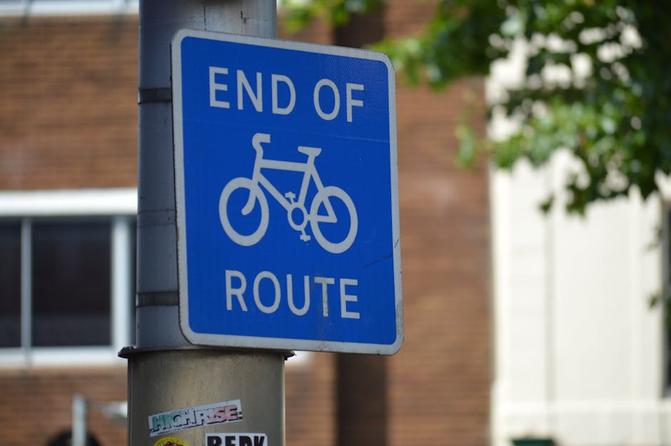 A blue rectangular sign mounted on a metal pole indicating the end of a bicycle route, with white text that reads 'END OF ROUTE' and a white bicycle symbol in the center. The sign is positioned outdoors in an urban environment, with a brick building and a window partially visible in the background. The pole has a small sticker attached near its base. The setting suggests a location where house removals or moving services by Man and Van Mile End may operate, with the sign situated on a street or pavement area close to residential buildings, supporting the context of home relocation logistics and transportation planning.