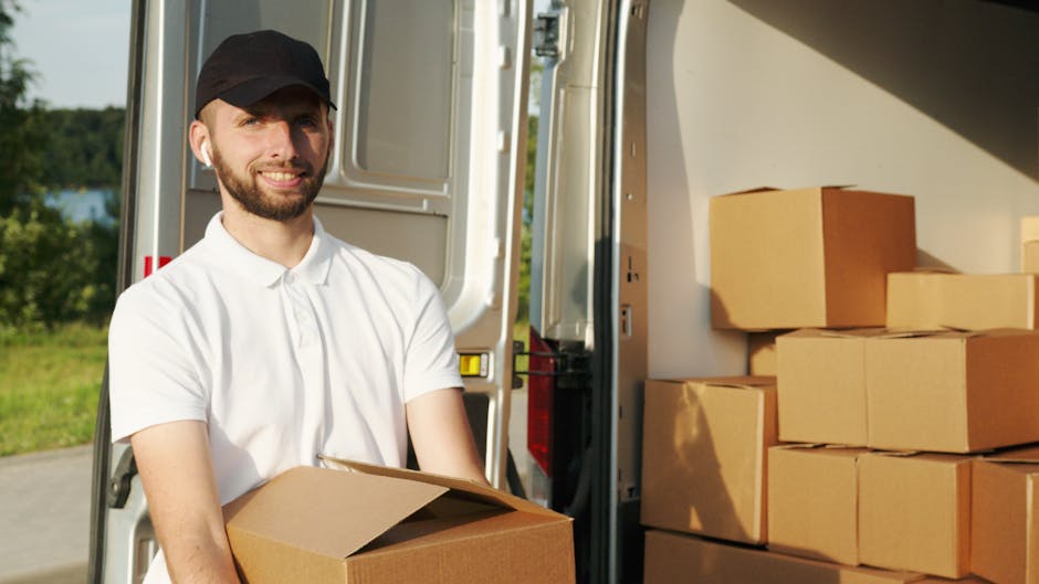 A professional mover from Man and Van Mile End is outdoors during a furniture transport and home relocation process, standing next to an open van filled with packed cardboard boxes and wrapped furniture items. The mover, wearing a white polo shirt and a black cap, is holding a filled cardboard box ready for loading or unloading. The van, parked on a paved area, has its back doors open, revealing the interior lined with additional boxes and packing materials. The background features green trees and a partly cloudy sky, indicating daytime lighting. The scene captures the loading process typical of house removals and packing and moving services, emphasizing careful handling of goods and organized logistics, aligned with the services offered by Man and Van Mile End.