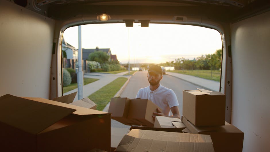 A man wearing a light-colored polo shirt and glasses is seen through the open rear door of a van, engaged in a home relocation process near Mile End Park during sunset. Inside the van, there are several cardboard boxes of varying sizes, some partially stacked and others ready for loading or unloading, with a few boxes placed on the van's floor. The man is handling a medium-sized box, appearing to either load it into the vehicle or prepare it for transport, with natural light illuminating the scene from outside. In the background, a quiet residential street with houses, greenery, and a body of water can be observed, highlighting the outdoor environment typical for local removals and furniture transport projects. Man and Van Mile End is involved in tasks such as packing, loading, and organizing belongings essential for efficient house moves, especially close to Mile End Park area.