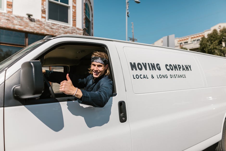 A professional mover from Man and Van Mile End is outdoors during a furniture transport and home relocation process, standing next to an open van filled with packed cardboard boxes and wrapped furniture items. The mover, wearing a white polo shirt and a black cap, is holding a filled cardboard box ready for loading or unloading. The van, parked on a paved area, has its back doors open, revealing the interior lined with additional boxes and packing materials. The background features green trees and a partly cloudy sky, indicating daytime lighting. The scene captures the loading process typical of house removals and packing and moving services, emphasizing careful handling of goods and organized logistics, aligned with the services offered by Man and Van Mile End.