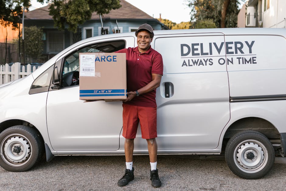 A man wearing a burgundy polo shirt, red shorts, black sneakers, and a grey cap is standing outside a white delivery van, holding a medium-sized cardboard box with a label and branding that reads 'LAR
