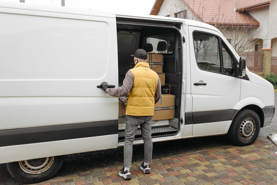 A man wearing a burgundy polo shirt, red shorts, black sneakers, and a grey cap is standing outside a white delivery van, holding a medium-sized cardboard box with a label and branding that reads 'LAR