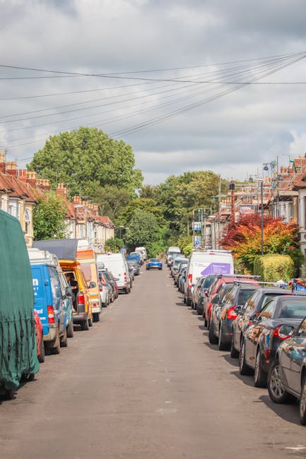 A residential street during daytime with parked cars lining both sides, including small vans and larger vehicles often used for house removals, some covered with protective tarps or blankets. The stre