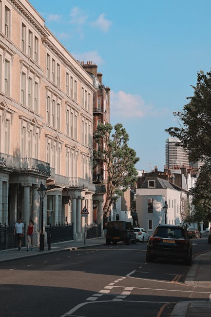 A residential street during daytime with parked cars lining both sides, including small vans and larger vehicles often used for house removals, some covered with protective tarps or blankets. The stre